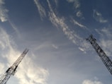 Two tall communication towers extend upwards against a backdrop of a clear sky. Wispy clouds are scattered across the blue expanse, creating a dynamic and open feel.