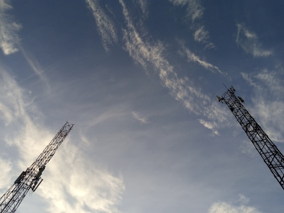 Two tall communication towers extend upwards against a backdrop of a clear sky. Wispy clouds are scattered across the blue expanse, creating a dynamic and open feel.