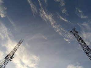 Two tall communication towers extend upwards against a backdrop of a clear sky. Wispy clouds are scattered across the blue expanse, creating a dynamic and open feel.