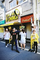A small group of people stands outside a shop called 'Lemonica.' The shop sign is bright yellow with a lemon illustration. The building is urban, with visible wires and windows above. A person in a yellow jacket, holding promotional material, is on the right.