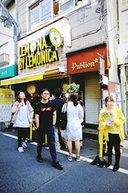 A small group of people stands outside a shop called 'Lemonica.' The shop sign is bright yellow with a lemon illustration. The building is urban, with visible wires and windows above. A person in a yellow jacket, holding promotional material, is on the right.