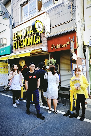 A small group of people stands outside a shop called 'Lemonica.' The shop sign is bright yellow with a lemon illustration. The building is urban, with visible wires and windows above. A person in a yellow jacket, holding promotional material, is on the right.