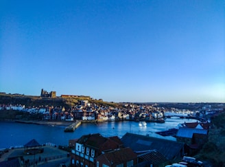 A coastal town with numerous old buildings sitting close to a large body of water under a clear blue sky. A ruined castle or abbey is visible atop a hill, overlooking the town. Brightly colored houses line the waterfront, and a dock area is visible with boats moored in the bay.