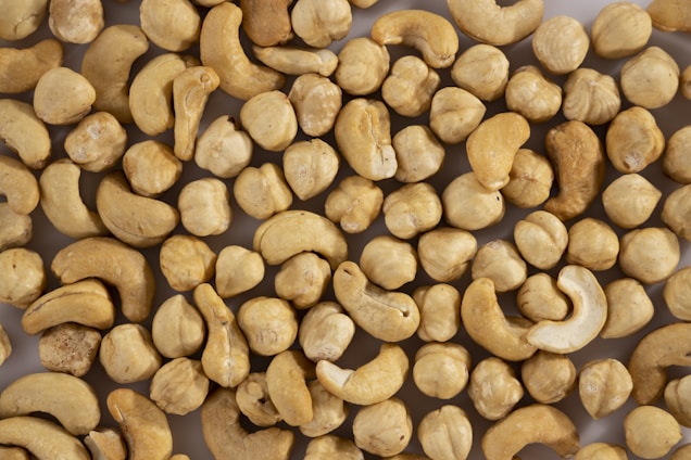 A collection of cashew nuts scattered across a light surface. The nuts vary slightly in shape and color, ranging from light beige to darker tan. Some nuts have a smooth surface, while others appear slightly rugged.