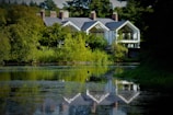 A serene landscape features a large, modern house nestled among lush greenery. The house boasts large glass windows and a reflective gray roof. A calm body of water in the foreground mirrors the building and surrounding trees, enhancing the tranquil atmosphere.
