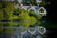 A serene landscape features a large, modern house nestled among lush greenery. The house boasts large glass windows and a reflective gray roof. A calm body of water in the foreground mirrors the building and surrounding trees, enhancing the tranquil atmosphere.