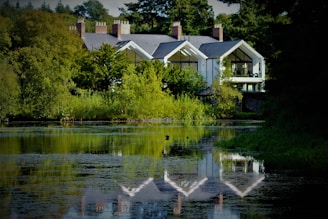 A serene landscape features a large, modern house nestled among lush greenery. The house boasts large glass windows and a reflective gray roof. A calm body of water in the foreground mirrors the building and surrounding trees, enhancing the tranquil atmosphere.