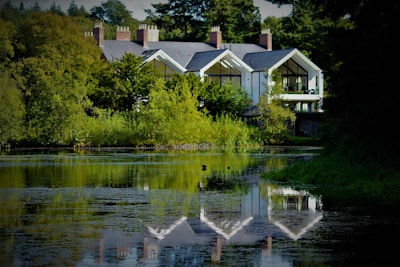 A serene landscape features a large, modern house nestled among lush greenery. The house boasts large glass windows and a reflective gray roof. A calm body of water in the foreground mirrors the building and surrounding trees, enhancing the tranquil atmosphere.