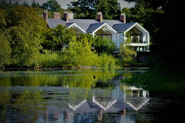 A serene landscape features a large, modern house nestled among lush greenery. The house boasts large glass windows and a reflective gray roof. A calm body of water in the foreground mirrors the building and surrounding trees, enhancing the tranquil atmosphere.
