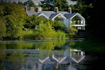 A serene landscape features a large, modern house nestled among lush greenery. The house boasts large glass windows and a reflective gray roof. A calm body of water in the foreground mirrors the building and surrounding trees, enhancing the tranquil atmosphere.