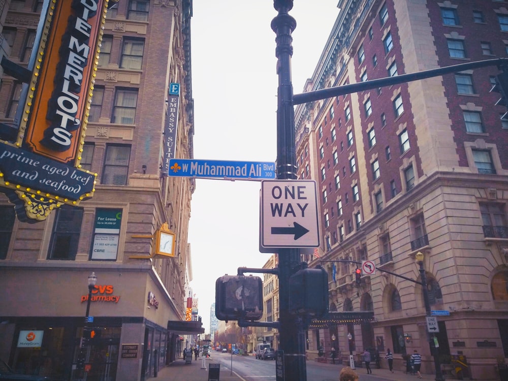 Image of Muhammad Ali Boulevard street sign in Louisville, KY. The sign is blue with white text and a yellow fleur-de-lis precedes the street name. There is also a one-way sign and crosswalk light on the street pole. Old brown buildings and businesses within them can be seen in the background.