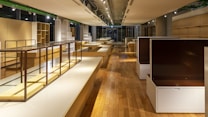 A spacious, well-lit interior with wooden floors and modern, minimalist shelving units. The shelves are empty, and the room is illuminated by overhead spotlights. The colors are warm, with a mix of natural wood tones and muted whites.