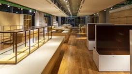 A spacious, well-lit interior with wooden floors and modern, minimalist shelving units. The shelves are empty, and the room is illuminated by overhead spotlights. The colors are warm, with a mix of natural wood tones and muted whites.