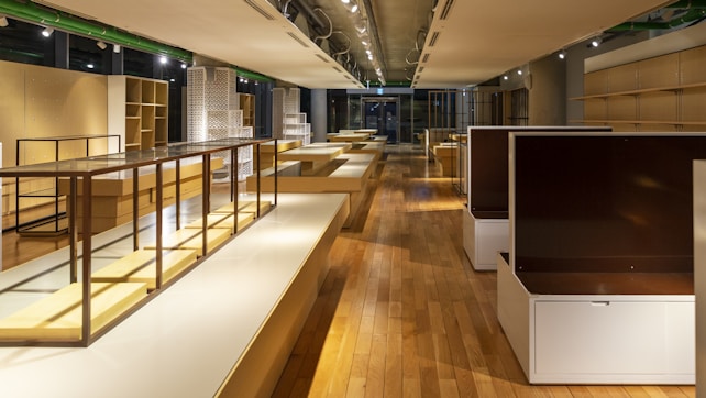 A spacious, well-lit interior with wooden floors and modern, minimalist shelving units. The shelves are empty, and the room is illuminated by overhead spotlights. The colors are warm, with a mix of natural wood tones and muted whites.