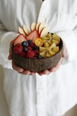 A person holds a coconut bowl filled with a variety of fresh, colorful fruits. The bowl contains sliced apples, strawberries, blueberries, raspberries, and starfruit, creating a visually appealing and vibrant arrangement.