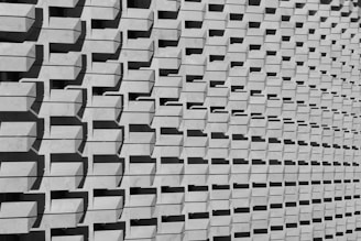 A repetitive pattern of rectangular concrete protrusions creates a textured, geometric facade. The parallel shadows cast by each protrusion add a rhythmic and dynamic visual element, enhancing the architectural interest of the structure.