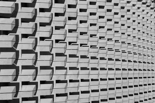 A repetitive pattern of rectangular concrete protrusions creates a textured, geometric facade. The parallel shadows cast by each protrusion add a rhythmic and dynamic visual element, enhancing the architectural interest of the structure.