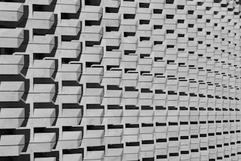 A repetitive pattern of rectangular concrete protrusions creates a textured, geometric facade. The parallel shadows cast by each protrusion add a rhythmic and dynamic visual element, enhancing the architectural interest of the structure.