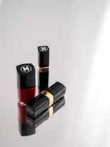 Three elegant cosmetic items, including two black lipstick tubes with gold bands and a bottle of red nail polish, arranged on a reflective surface. The items display a luxurious brand logo on a minimalist grey background.