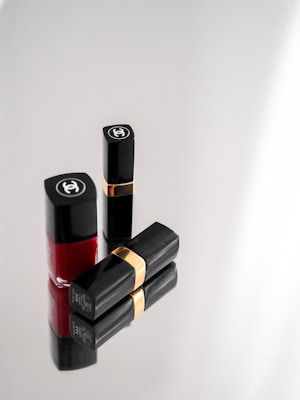 Three elegant cosmetic items, including two black lipstick tubes with gold bands and a bottle of red nail polish, arranged on a reflective surface. The items display a luxurious brand logo on a minimalist grey background.