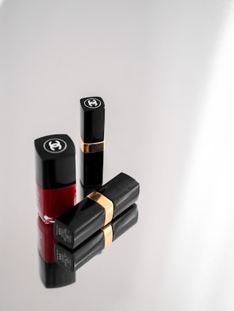 Three elegant cosmetic items, including two black lipstick tubes with gold bands and a bottle of red nail polish, arranged on a reflective surface. The items display a luxurious brand logo on a minimalist grey background.