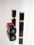 Three luxury cosmetic containers are placed on a reflective surface. Two of them are upright with black and gold detailing, while one is lying down showcasing a bold red color with a logo clearly visible.