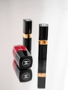 Three luxury cosmetic containers are placed on a reflective surface. Two of them are upright with black and gold detailing, while one is lying down showcasing a bold red color with a logo clearly visible.