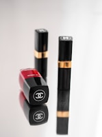 Three luxury cosmetic containers are placed on a reflective surface. Two of them are upright with black and gold detailing, while one is lying down showcasing a bold red color with a logo clearly visible.