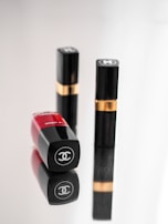 Three luxury cosmetic containers are placed on a reflective surface. Two of them are upright with black and gold detailing, while one is lying down showcasing a bold red color with a logo clearly visible.