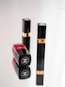Three luxury cosmetic containers are placed on a reflective surface. Two of them are upright with black and gold detailing, while one is lying down showcasing a bold red color with a logo clearly visible.