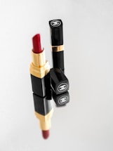 A luxurious black and gold lipstick case with a red lipstick bullet partially extended. Next to it are two closed lipstick cases, all displaying a prominent luxury brand logo on the top. The arrangement is reflected on a glossy surface.