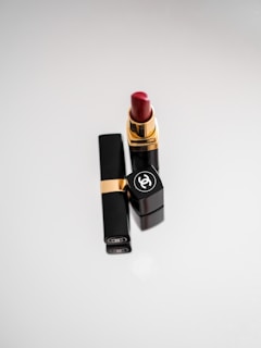 A tube of luxurious red lipstick stands upright, with an elegant black casing featuring a gold accent and a prominent logo on the cap. The background is a soft, light gradient that adds a sophisticated touch.