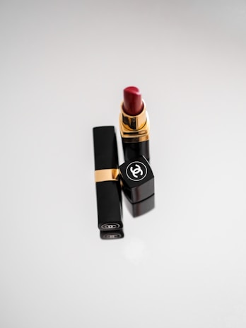 A tube of luxurious red lipstick stands upright, with an elegant black casing featuring a gold accent and a prominent logo on the cap. The background is a soft, light gradient that adds a sophisticated touch.