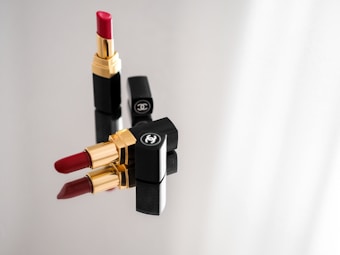 Three lipsticks with golden and black casings are arranged on a reflective surface. Two lipsticks are closed with their caps on, while one is uncapped revealing red lipstick. The brand logo is visible on the caps.
