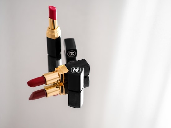 Three lipsticks with golden and black casings are arranged on a reflective surface. Two lipsticks are closed with their caps on, while one is uncapped revealing red lipstick. The brand logo is visible on the caps.