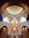 A grand chandelier hangs from an ornate ceiling, featuring intricate geometric and floral patterns. The walls are adorned with detailed carvings and Arabic script, enhancing the luxurious and elegant ambiance of the space.