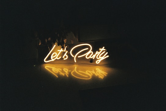 A neon sign displaying the words 'Let's Party' in bright, glowing yellow with a reflection on a glossy surface below. The background is mostly dark, enhancing the vibrancy of the sign.