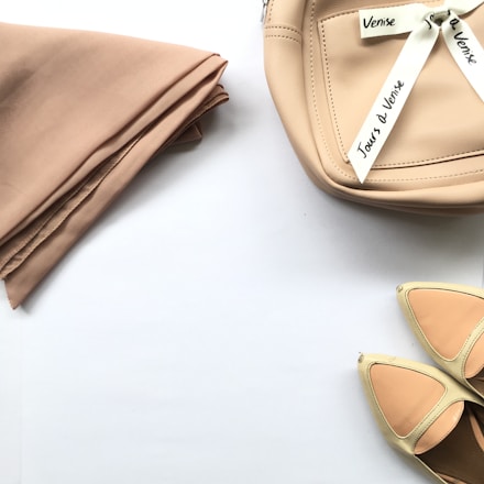 A neatly folded beige cloth placed on a white surface, accompanied by a beige leather bag adorned with white ribbons that have text written on them. Near the bag is a pair of shoes featuring a combination of cream and peach shades.