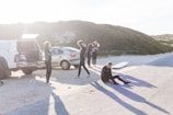 Several people in wetsuits are gathered near two parked vehicles. One person is sitting on the ground while others appear to be preparing or conversing. A surfboard is lying on the sand nearby, and the scene is lit by bright sunlight casting long shadows.