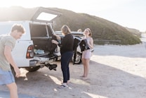 A group of people stands near a white vehicle on a sandy area with greenery in the background. The vehicle's back is open, showing various items, and a surfboard is visible next to the car. One person is holding a wetsuit, and another is adjusting something in the back of the car.