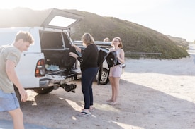 A group of people stands near a white vehicle on a sandy area with greenery in the background. The vehicle's back is open, showing various items, and a surfboard is visible next to the car. One person is holding a wetsuit, and another is adjusting something in the back of the car.