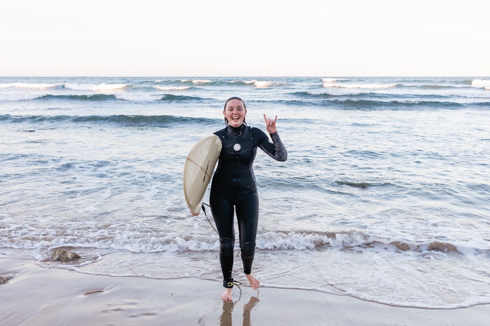 Mujer con traje de neopreno negro sosteniendo una tabla de surf blanca de pie en la playa durante el día