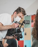 A person wearing a white face mask is closely operating a professional video camera, focusing intently while capturing footage. The background features colorful lockers in shades of blue, red, and beige.