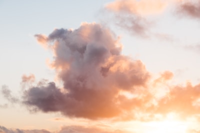 A large cloud formation dominates the sky, tinged with warm colors from the setting sun. The clouds appear fluffy and dense, with gentle hues of pink, orange, and purple. The sky in the background is mostly clear, with a subtle gradient from light blue to soft yellow near the horizon.