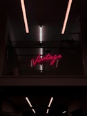Neon pink sign displaying the word 'Vintage' placed on a dark, industrial-style wall with metal mesh. Overhead, linear fluorescent lights cast a soft glow.
