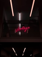 Neon pink sign displaying the word 'Vintage' placed on a dark, industrial-style wall with metal mesh. Overhead, linear fluorescent lights cast a soft glow.