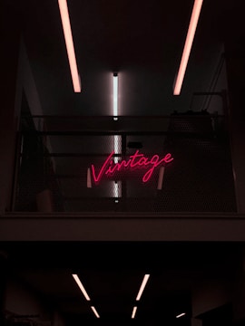 Neon pink sign displaying the word 'Vintage' placed on a dark, industrial-style wall with metal mesh. Overhead, linear fluorescent lights cast a soft glow.