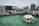 A rooftop area with green artificial grass flooring and a stack of white chairs arranged upside down in a circular formation. The background features a city skyline with many tall buildings under an overcast sky.