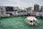 A rooftop area with green artificial grass flooring and a stack of white chairs arranged upside down in a circular formation. The background features a city skyline with many tall buildings under an overcast sky.