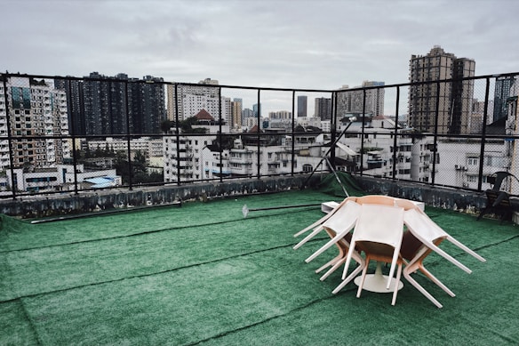 A rooftop area with green artificial grass flooring and a stack of white chairs arranged upside down in a circular formation. The background features a city skyline with many tall buildings under an overcast sky.